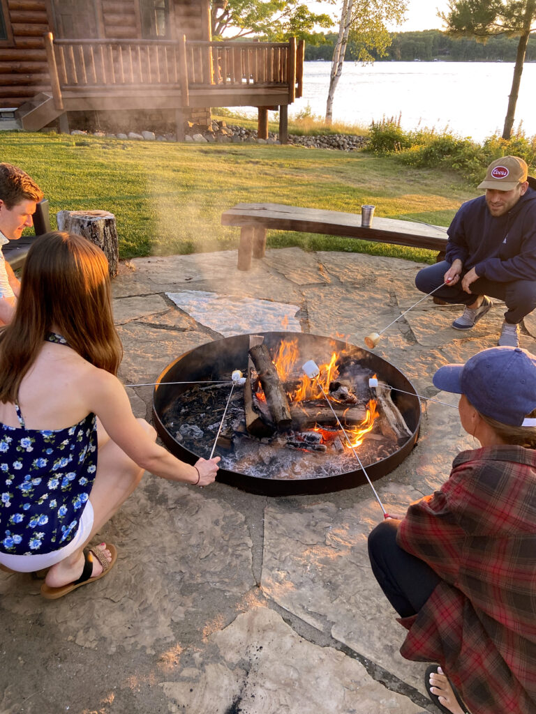 Four people crouching next to a fire pit roasting marshmallows.