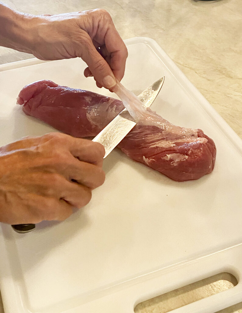 Person with a knife cutting off the silver skin membrane from a pork tenderloin.