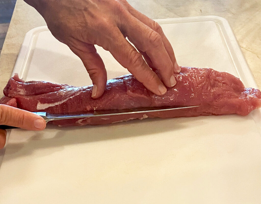 Person with a knife cutting a lengthwise slit into a pork tenderloin.