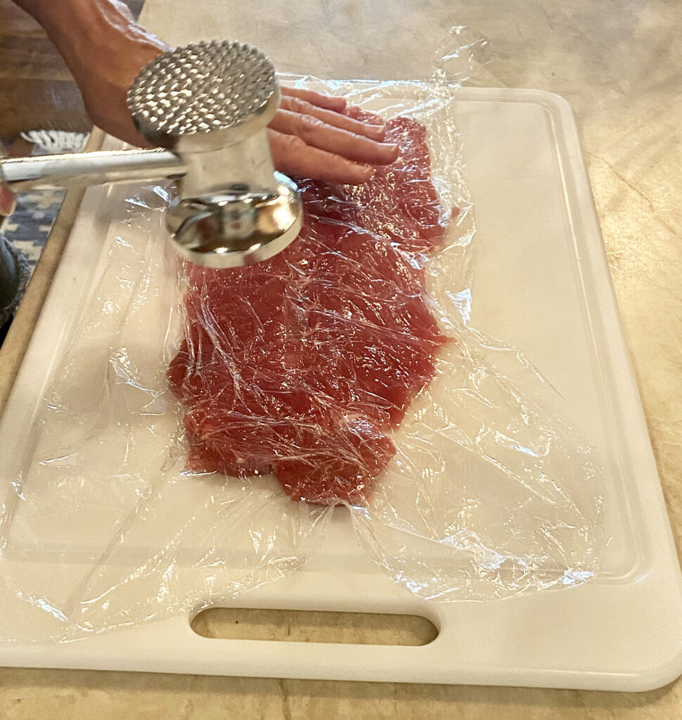 Person with a meat mallet pounding a split pork tenderloin flat.