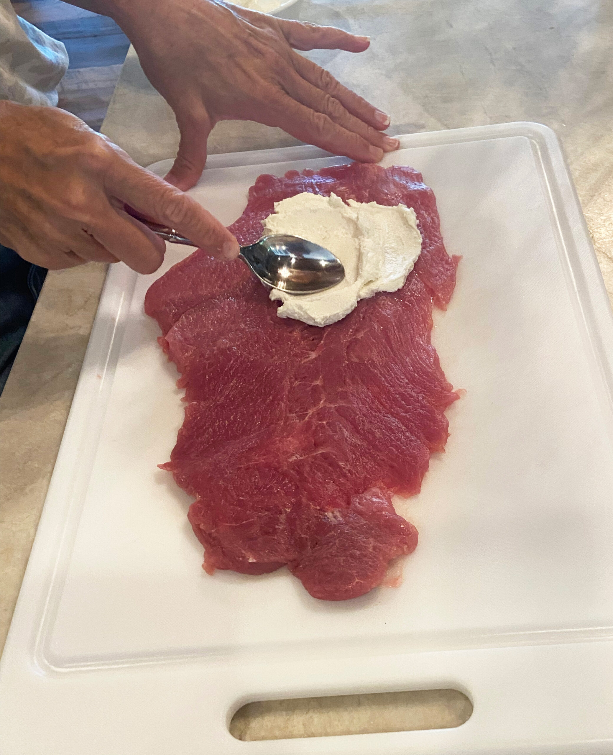 Person using a spoon to spread goat cheese over the surface of a flattened pork tenderloin.